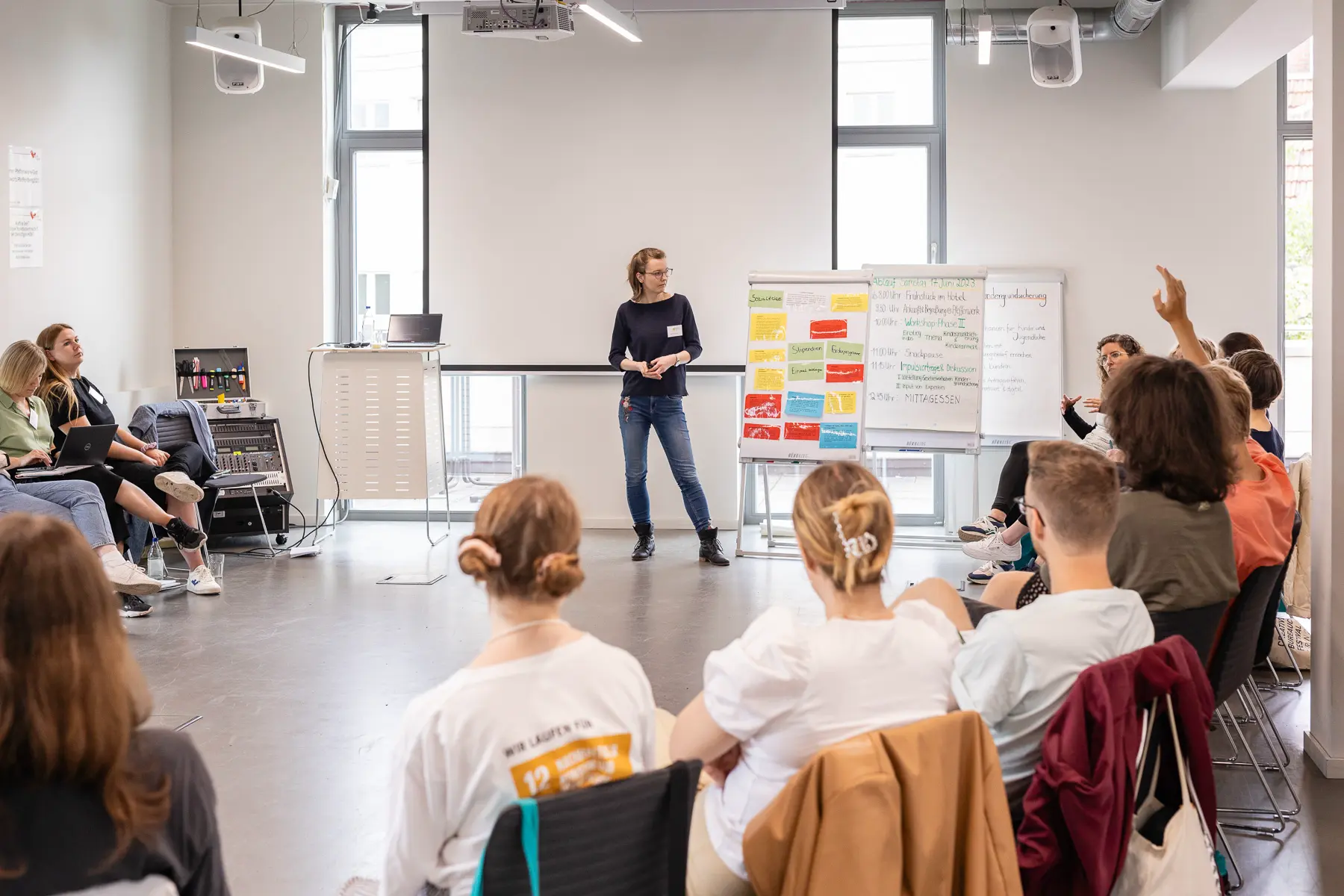 Gruppendiskussion im Seminarraum mit einer Moderatorin neben drei Flipcharts. Die Teilnehmenden sitzen im Stuhlkreis, ein Teilnehmer meldet sich.