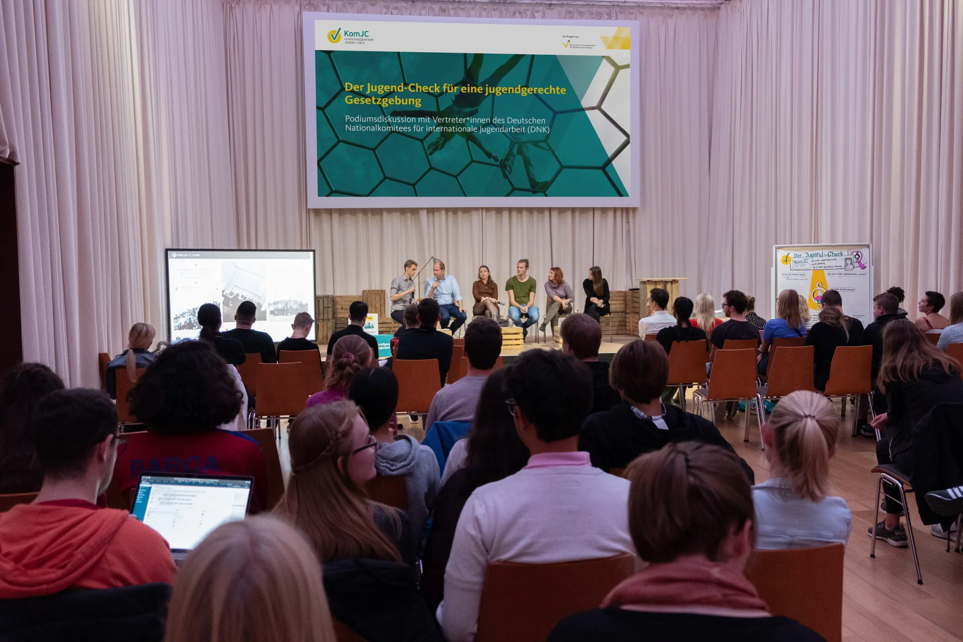 Blick in einen gefüllten Veranstaltungsraum mit einer Podiumsdiskussion im Hintergrund. Auf dem Podium waren (v.l.n.r.): Bernd Fiedler, Jan Holze, Susanne Zels, Georg Kurz, Ana-Maria Trăsnea und Laura Schieritz.