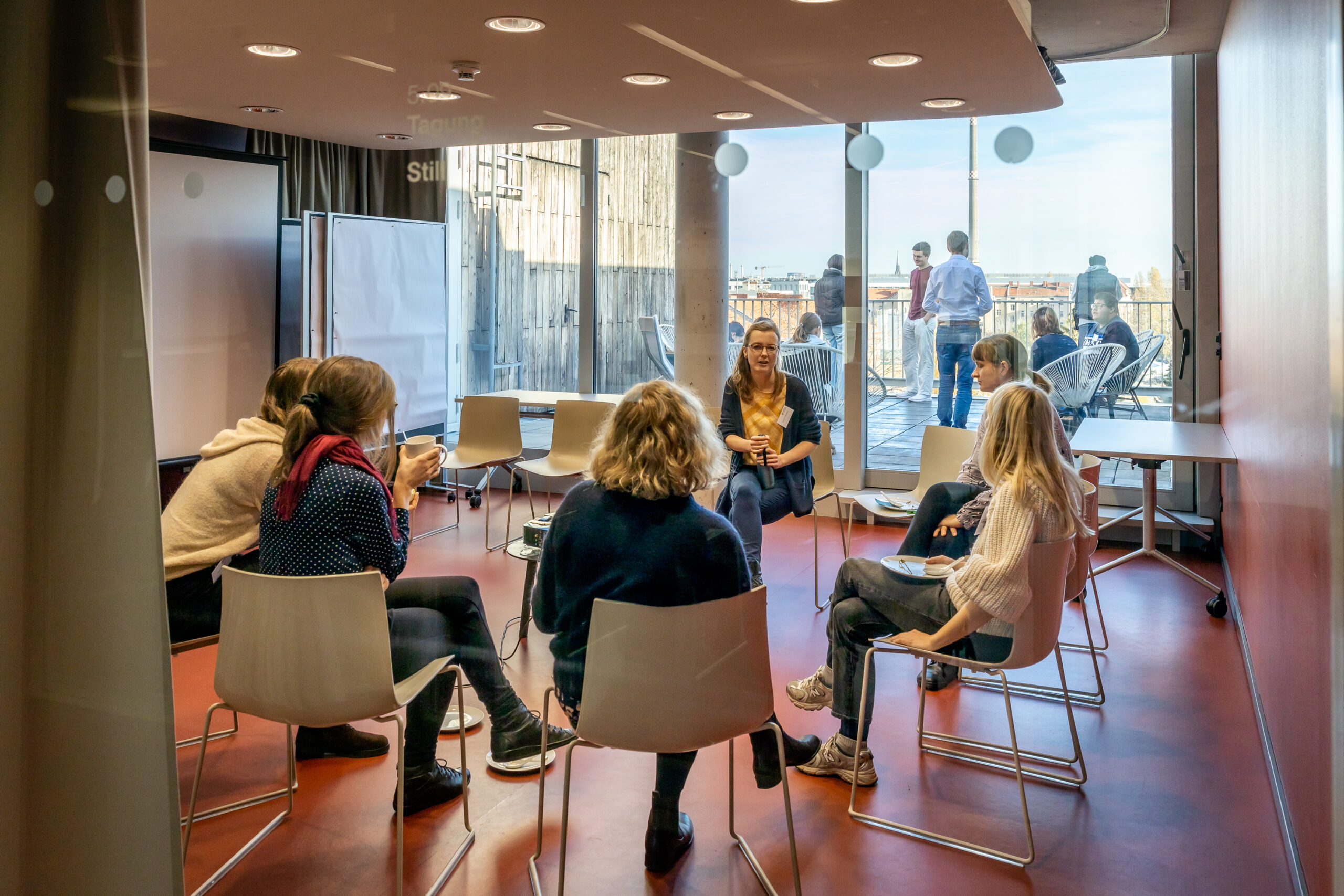 Seminarraum mit Gruppe von jungen Menschen im Stuhlkreis, im Hintergrund Dachterrasse mit jungen Menschen und Blick über Berlin.