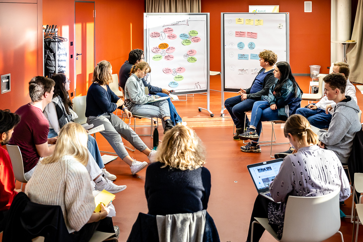 Junge Menschen diskutieren im Stuhlkreis, im Hintergrund zwei beschriebene Pinnwände.