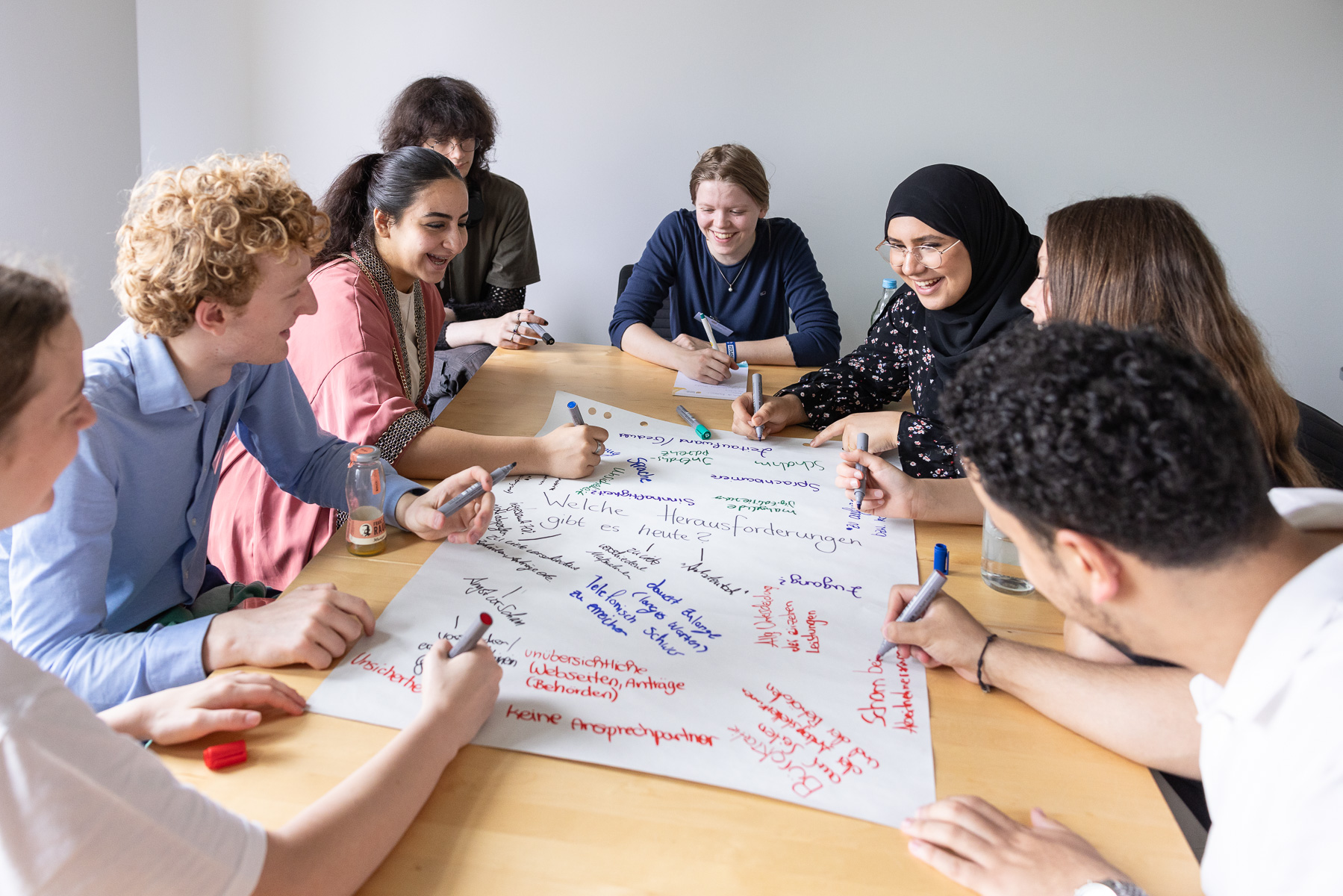 Eine Gruppe junger Menschen, die im Rahmen eines Workshops gemeinsam an einem Flipchart-Papier arbeiten und sich dabei lachend unterhalten.