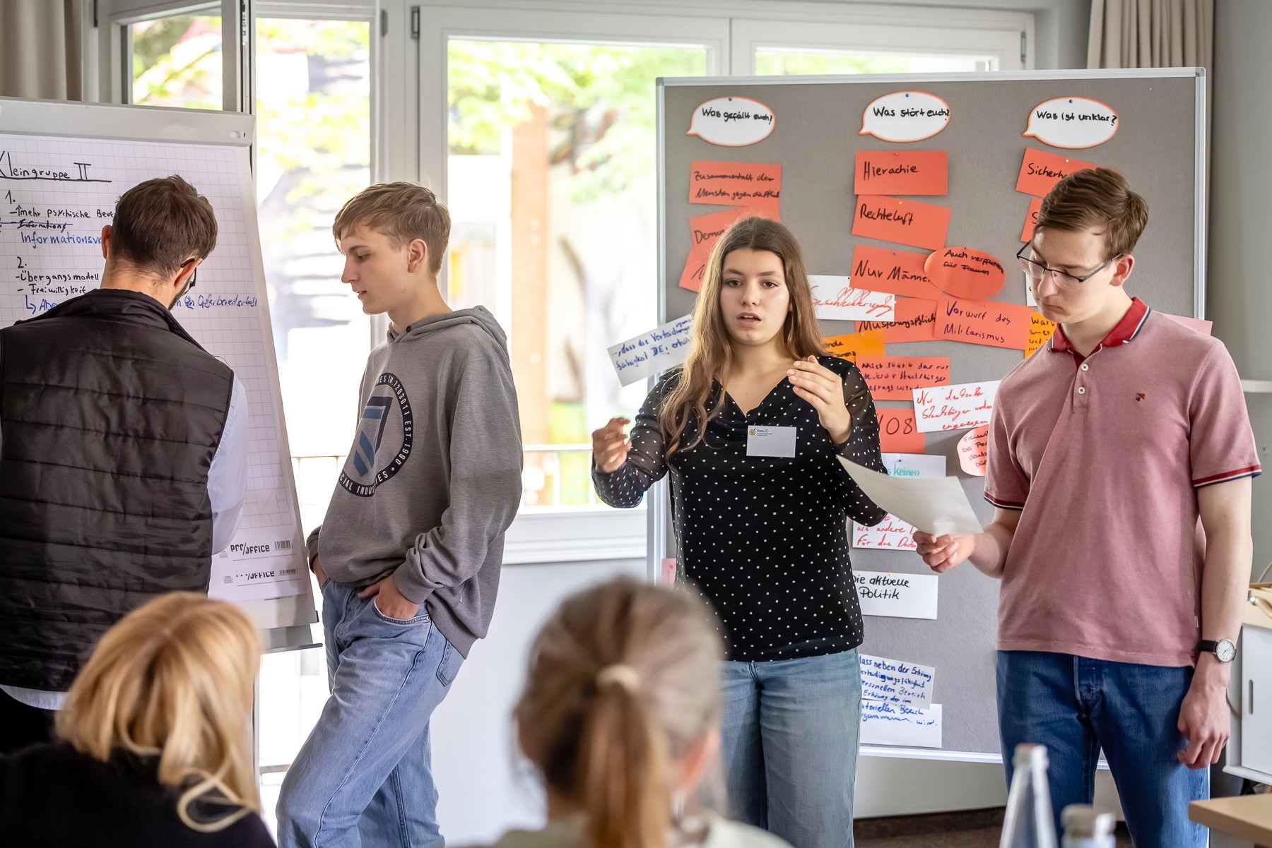 Mehrere Teilnehmende in einer Kleingruppe präsentieren ihre Ergebnisse. Sie stehen vor Pinnwänden mit beschrifteten Zetteln.